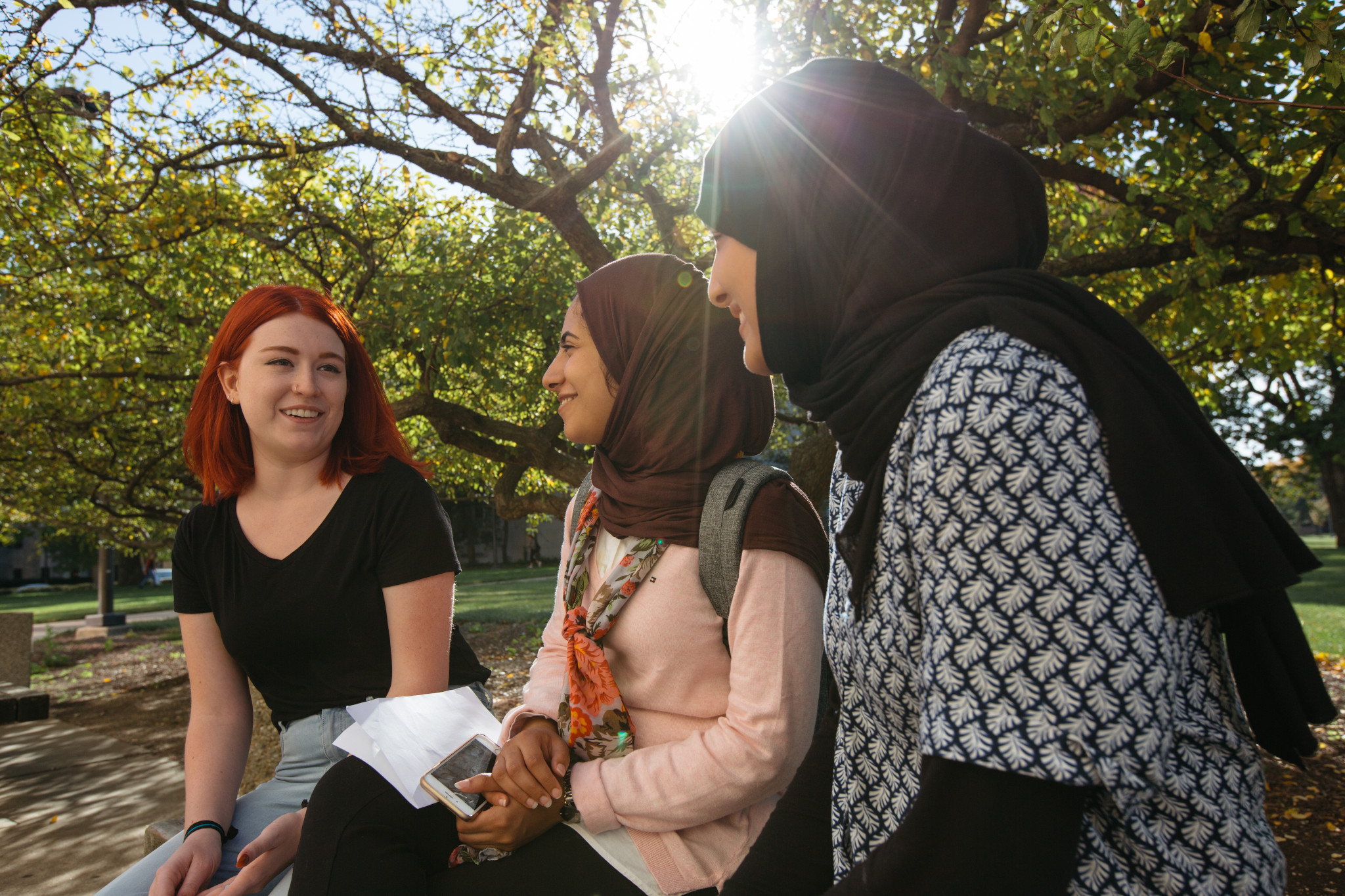 A group of three women sit on a bench outdoors talking to eachother